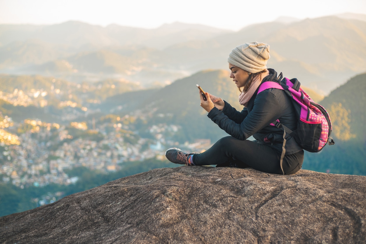 Femme assise sur un rocher en haut d'un point panoramique qui regarde son téléphone