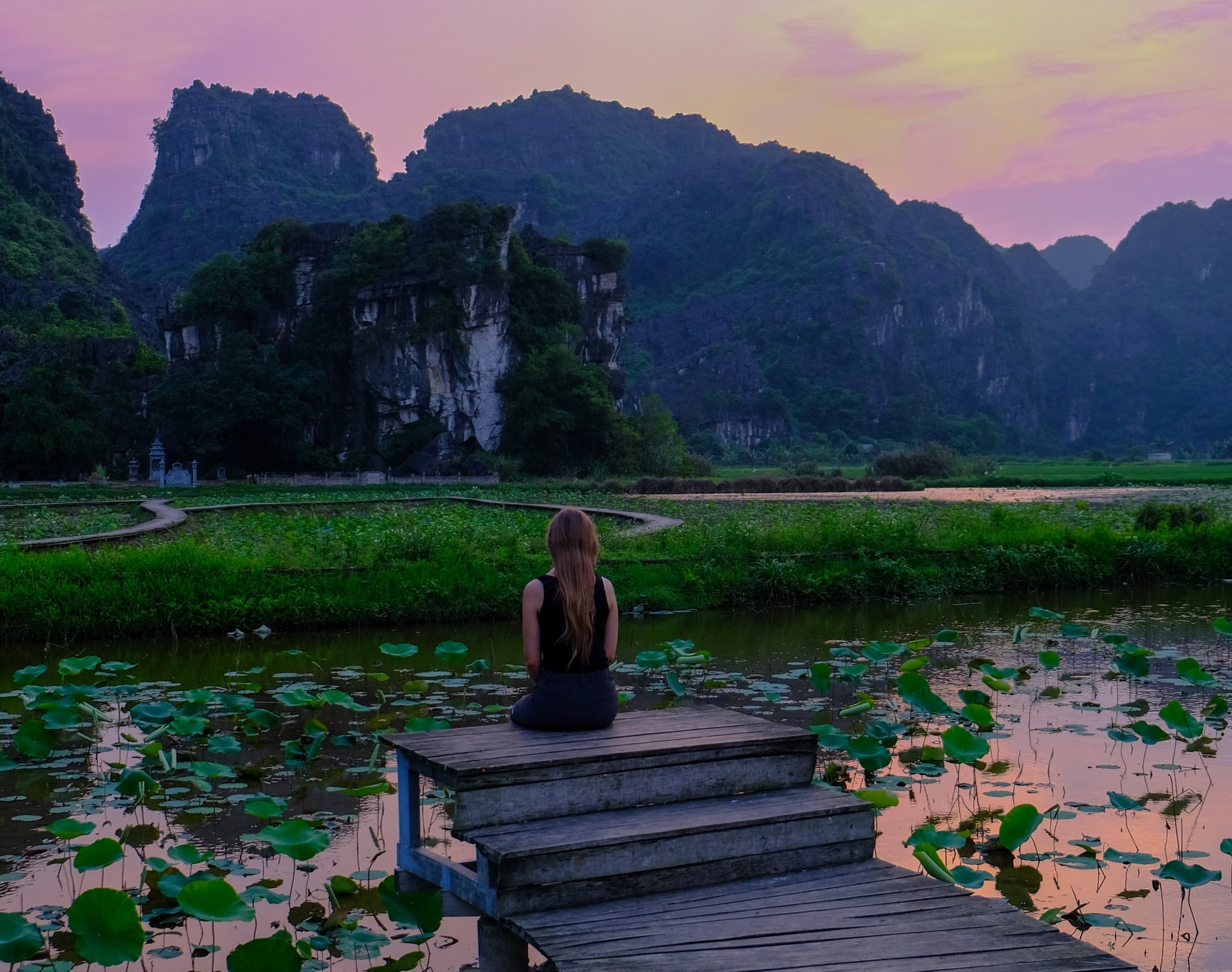 jeune femme face aux montagnes devant une étendue d'eau au coucher du soleil