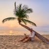 Jeune homme travaillant sur son ordinateur portable sous un palmier sur une plage au coucher du soleil