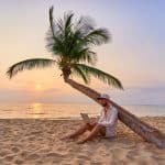 Jeune homme travaillant sur son ordinateur portable sous un palmier sur une plage au coucher du soleil