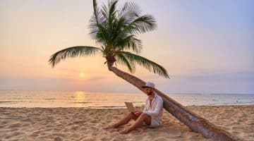 Jeune homme travaillant sur son ordinateur portable sous un palmier sur une plage au coucher du soleil