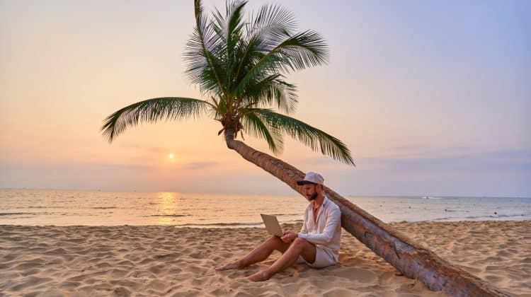 Jeune homme travaillant sur son ordinateur portable sous un palmier sur une plage au coucher du soleil