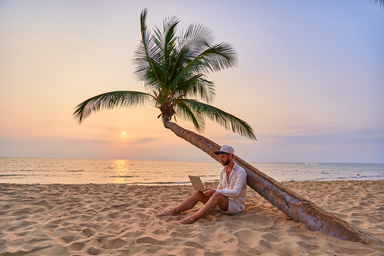 Jeune homme travaillant sur son ordinateur portable sous un palmier sur une plage au coucher du soleil
