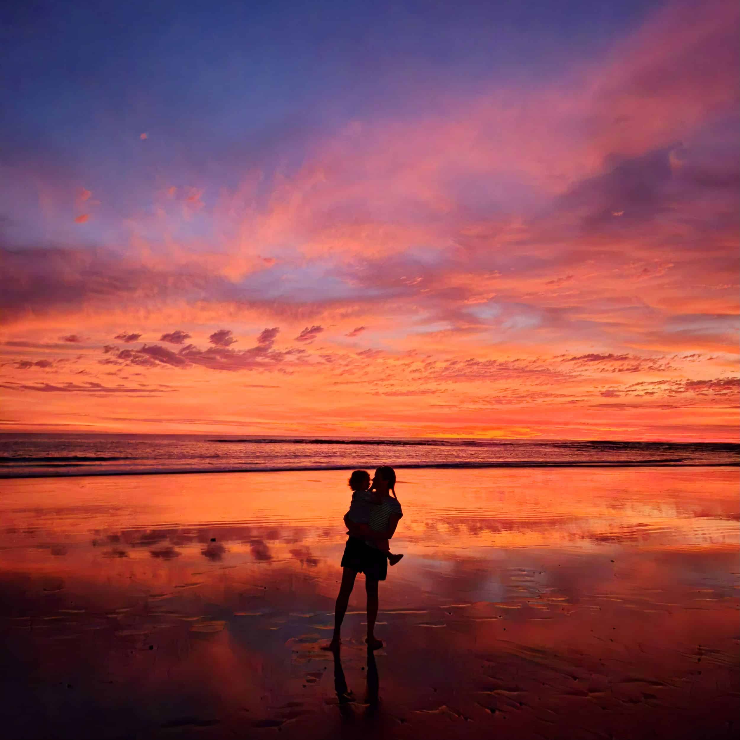 Mère portant sa fille au bord de l'eau sur la plage au coucher du soleil