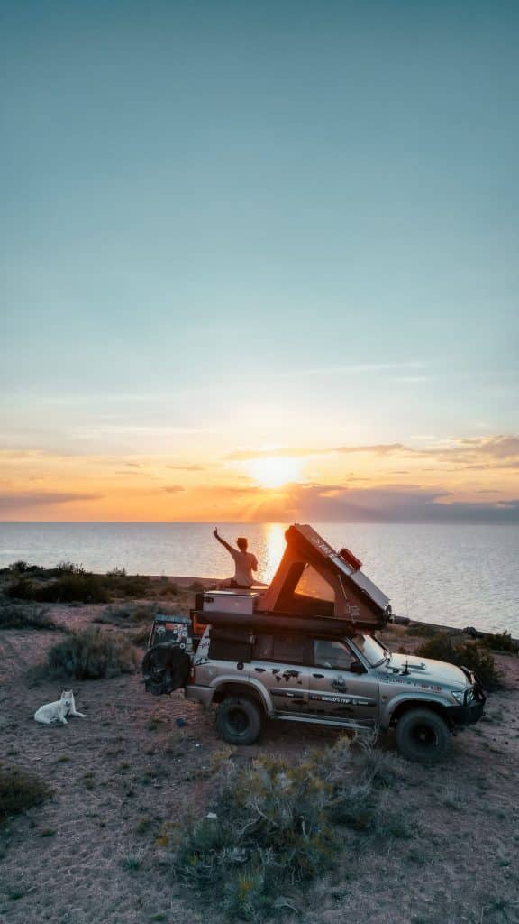 Jeune femme sur le toit de son 4x4 levant les bras devant le coucher de soleil